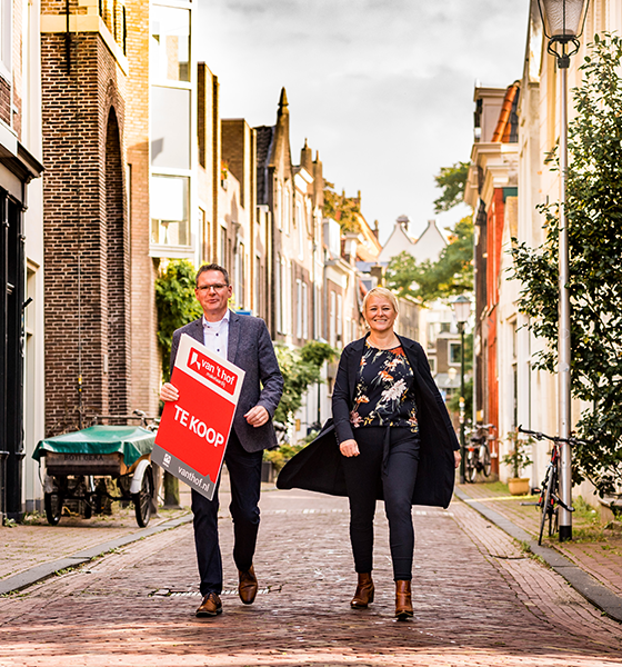 Twee makelaars die naar de camera wandelen met een verkoopbord in handen.