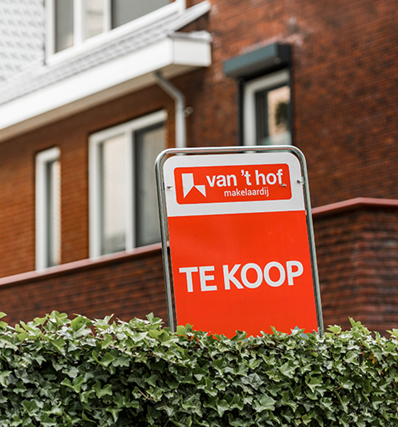 Verkoopbord van van 't Hof Makelaardij in de heg van een te-koop staande woning
